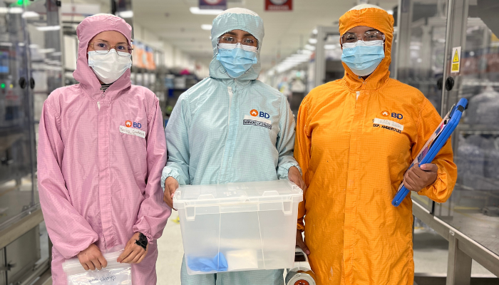 Tres asociados de BD con vestimenta de sala limpia sosteniendo un contenedor de almacenamiento.