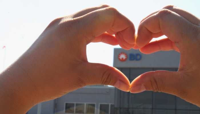 Manos formando la figura de un coraz&oacute;n con un edificio de BD al fondo.