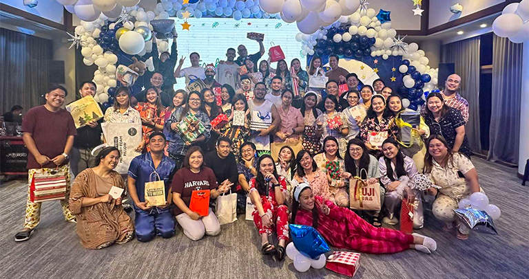 Large group gathered in a decorated event space featuring an arch of white and blue balloons, star accents, and a screen backdrop, with many participants holding wrapped gifts and posing together for a group photo.