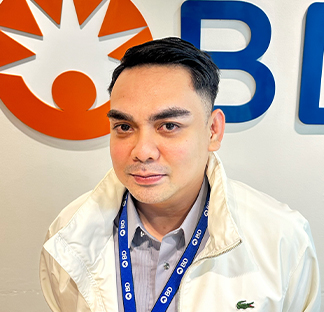Individual wearing a light-colored collared shirt and a white jacket, standing indoors in front of a wall displaying a large BD logo with orange and blue lettering. A lanyard with a repeating BD design hangs around the individual's neck.