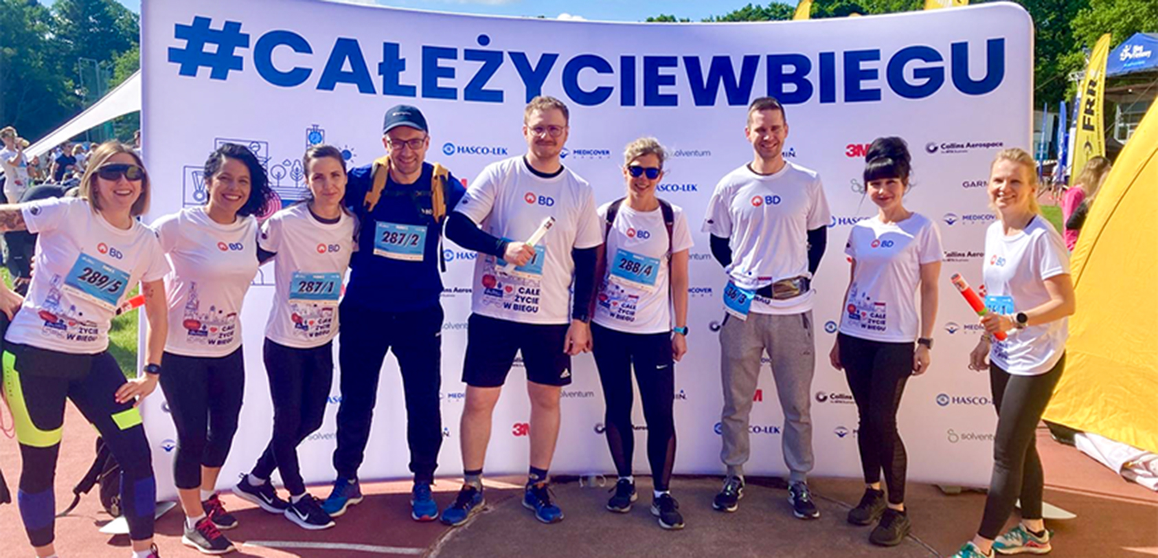 Group of nine people wearing athletic attire standing outside in front of a white cloth backdrop