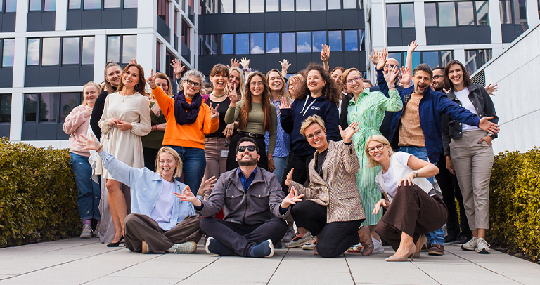 A wide outdoor group photo of individuals posing in front of a modern office building with reflective glass panels. The group is positioned on a paved terrace bordered by hedges, with people standing and sitting in several rows.