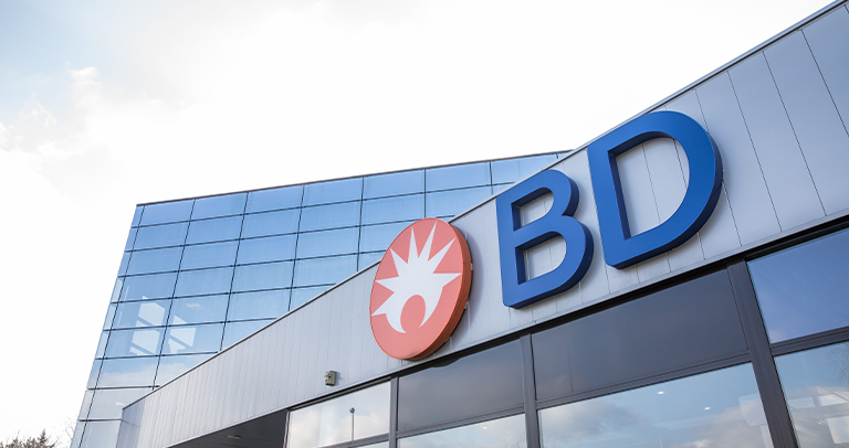 Exterior view of a modern BD office building with reflective glass windows and a large BD logo mounted on the front facade, photographed under a bright sky.