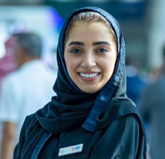 Individual wearing a dark-colored garment and a head covering, standing indoors with a blurred background of people and displays. A BD-branded name badge is attached to the garment.