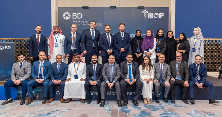 Large group of individuals posed indoors in front of a backdrop displaying the BD logo, the phrase ‘Advancing the world of health,’ and event branding. The group is arranged in two rows, with some seated in front and others standing behind on a stage-like