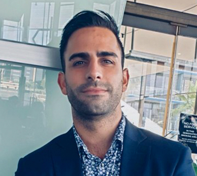 Individual in a portrait-style image wearing a dark blazer and patterned shirt, standing indoors near a glass wall with city buildings visible in the background.