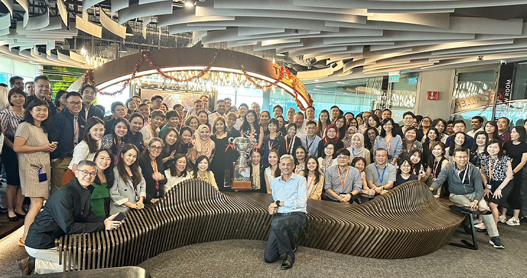 Wide-angle group photo of individuals gathered inside a modern indoor space featuring sculptural ceiling panels and a curved wooden seating structure. The group is arranged around a large decorative trophy displayed at the center.