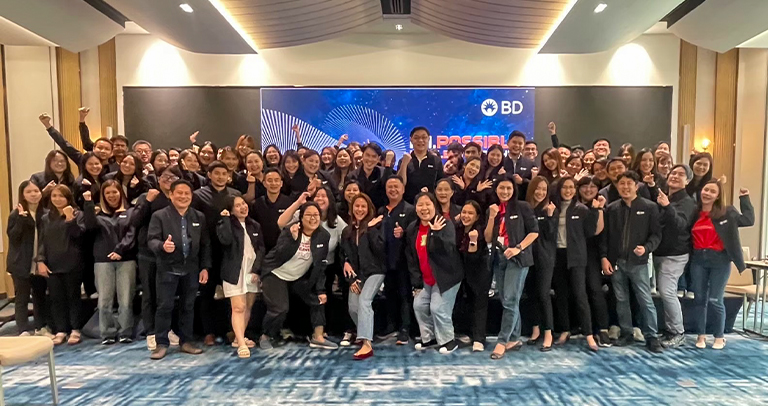 Large group of individuals standing together in a conference room in front of a BD-branded digital screen displaying abstract blue graphics.