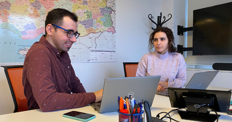 Two people are seated at a desk with laptops, engaged in conversation. A map is visible in the background, creating a professional office setting.