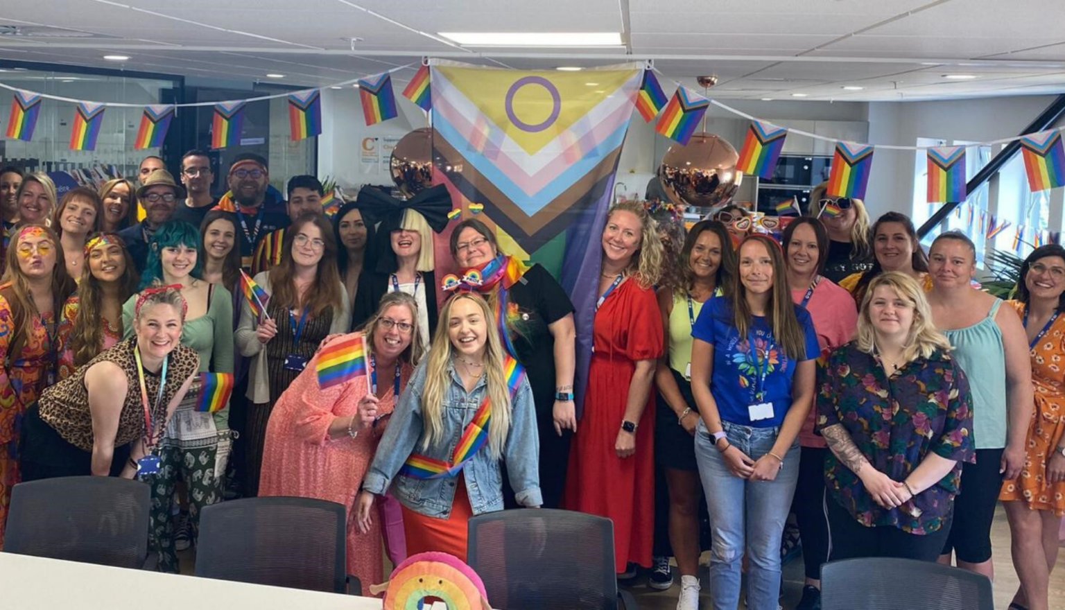Large group of BD associates gathered indoors holding Pride flags at a workplace event.