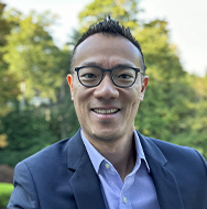 Individual wearing glasses and a dark blazer over a light collared shirt, photographed outdoors with green trees softly blurred in the background.