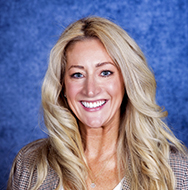 Individual with long light-colored hair wearing a patterned top, photographed in a close-up portrait against a blue textured background.