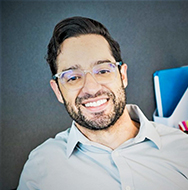 Individual wearing glasses and a light-colored collared shirt seated indoors with a dark wall and brightly colored folders in the background.