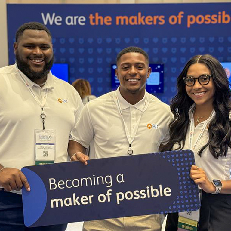 Three people smiling and holding a sign that reads "Becoming a maker of possible." They wear white shirts with a BD logo. Background includes a blue banner.