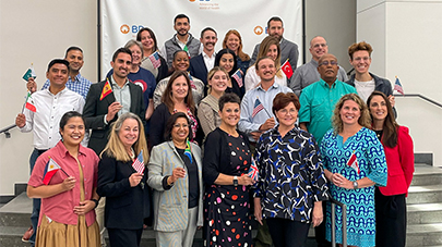 A diverse group of smiling people stand on steps, holding various national flags. They are dressed in business and casual attire, projecting a joyful and inclusive atmosphere.