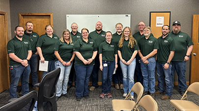 A group of 14 people in green polo shirts and blue jeans stand in a classroom. They are smiling, suggesting a friendly, team-oriented atmosphere.