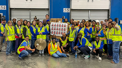 A diverse group of smiling warehouse workers in yellow safety vests pose together, holding a board with badges. The setting is a large industrial space.