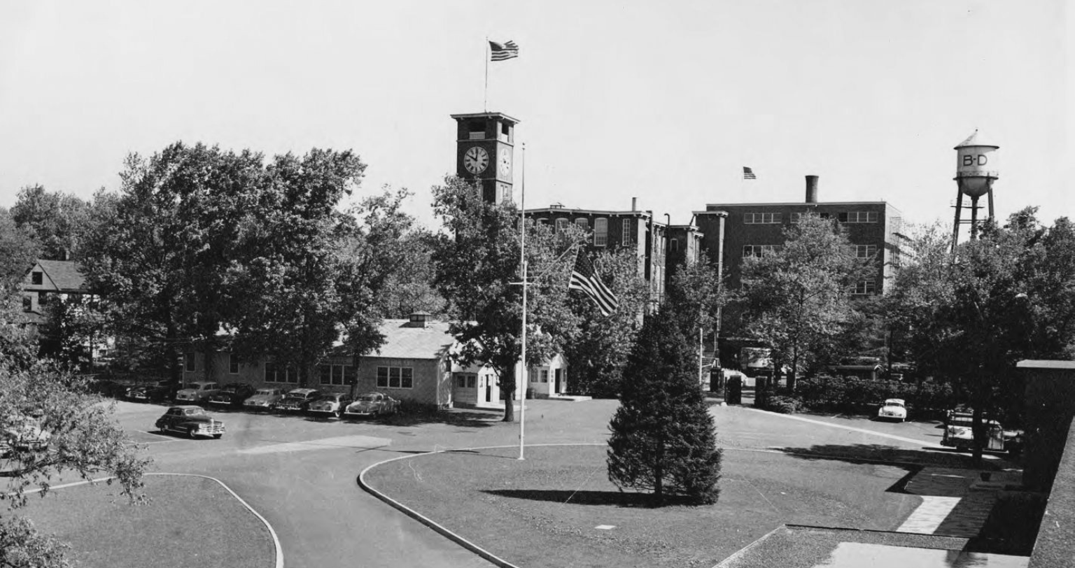 Historic black‑and‑white photo of a BD facility with a central clock tower and surrounding buildings.