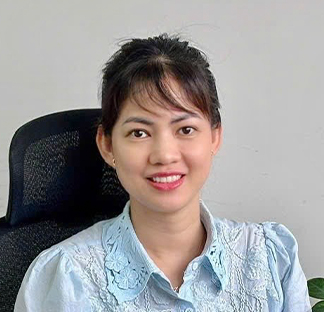 Individual seated in an office chair in front of a plain light‑colored wall, wearing a light blue embroidered top. The person is positioned close to the camera in a portrait‑style photo with part of a black chair visible behind.