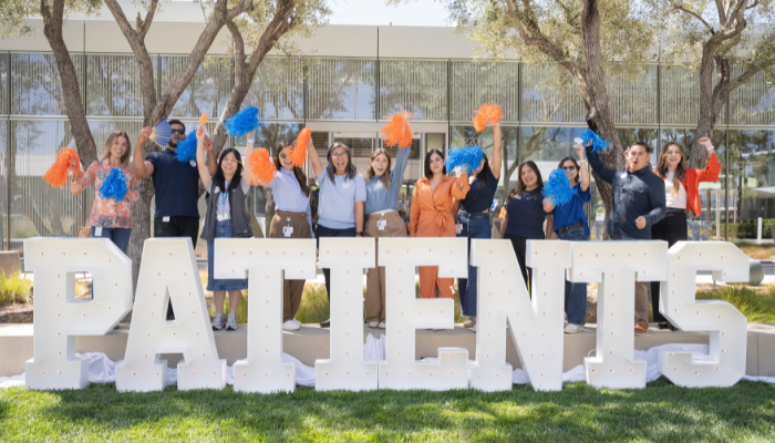 Grupo de associados da BD em &aacute;rea externa, posicionados atr&aacute;s de grandes letras com a palavra &ldquo;PATIENTS&rdquo; e segurando pompons azuis e laranja.