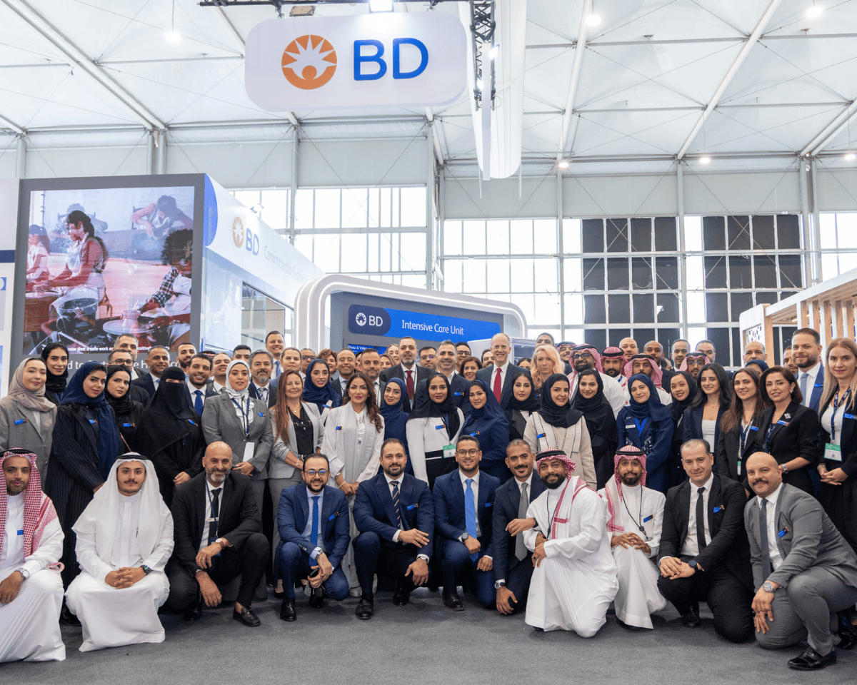 Grande grupo de associados da BD reunidos em frente ao estande da BD em um evento, com a marca BD visível ao fundo.