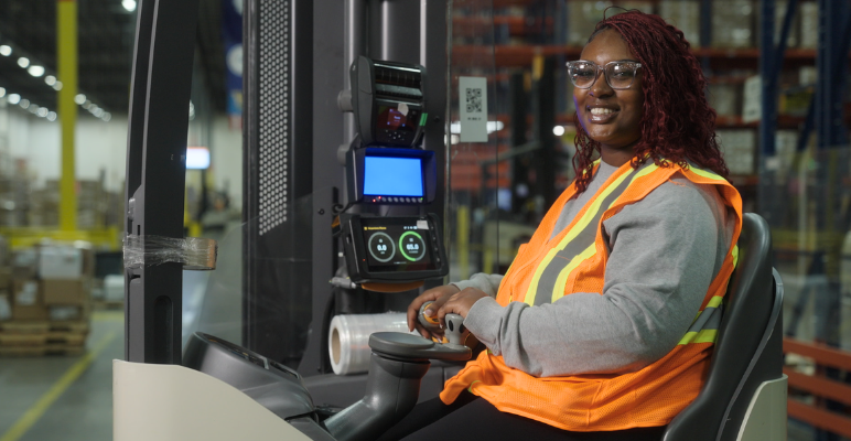 Image showing a person in a high‑visibility safety vest operating powered warehouse equipment inside a distribution facility with aisles of shelving and stacked materials.