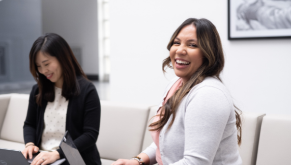Una persona sonriendo a la c&aacute;mara, mientras otra trabaja en su port&aacute;til en segundo plano.