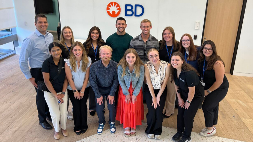 Un gran grupo de asociados sonriendo juntos frente a una pared blanca con el logotipo de BD.