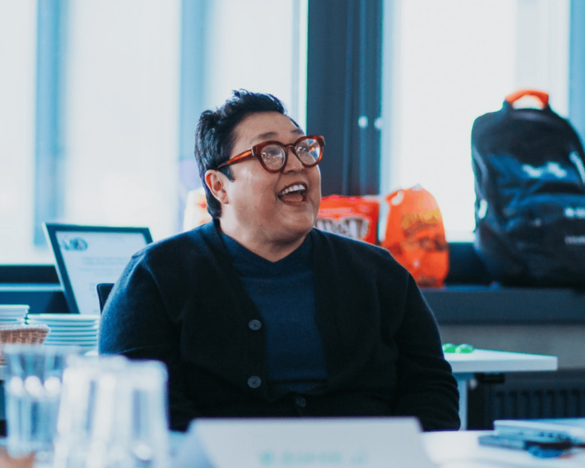 Una persona de cabello corto y oscuro, sonriendo y conversando. La persona lleva gafas y un suéter oscuro en un entorno corporativo.