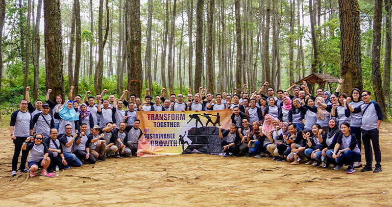 Large group of individuals gathered outdoors in a forested area, standing behind a banner that reads ‘Transform Together for Sustainable Growth.’ Tall trees surround the group on all sides, and the ground is covered in packed dirt.