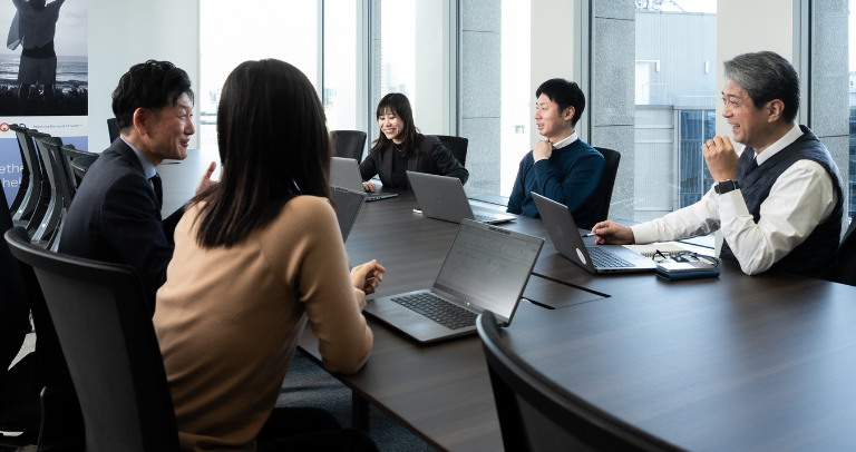 屋内の長方形のテーブルを囲んで5人が着席し、それぞれがノートパソコンを開いて互いに向き合っている。背景には大きな窓が広がる、会議の場面。