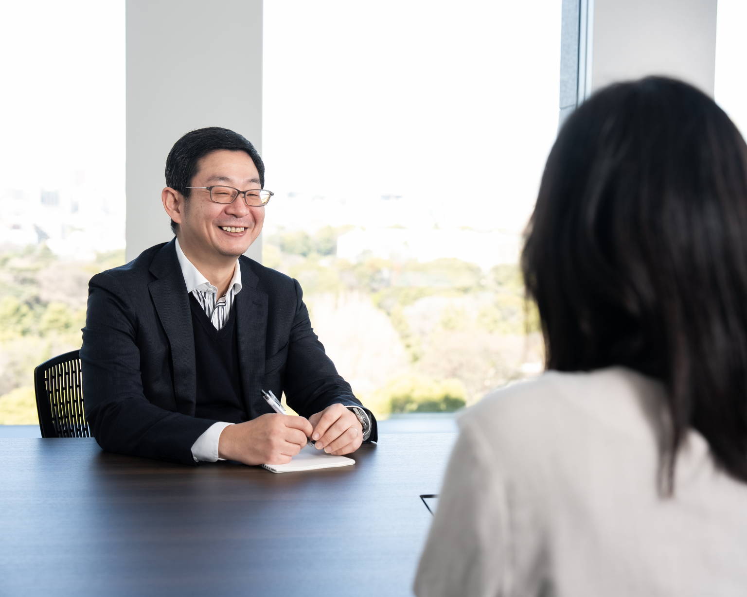 屋内のテーブルに座った人物がペンを手にし、手前に背中が見えている別の人物と向かい合っている。背景には窓が写っている。