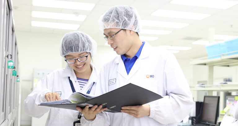 白衣、ヘアカバー、眼鏡を着用した2人が屋内に立ち、開いたバインダーか書類を二人で覗き込んでいる。