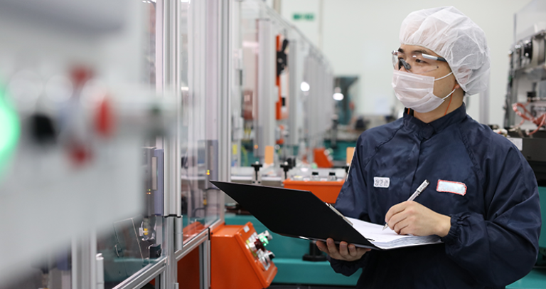 屋内にて、顔用マスク、頭部カバー、眼鏡、および防護服を着用した人物が、密閉された機械の横に立ち、クリップボードとペンを手にしている。