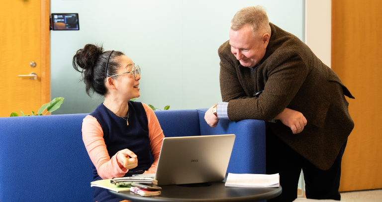 屋内の小さなテーブルのそばで、座ったり立ったりしている2人が、開いたノートパソコンやノート、私物などが見える中、互いに向き合って会話をしている。