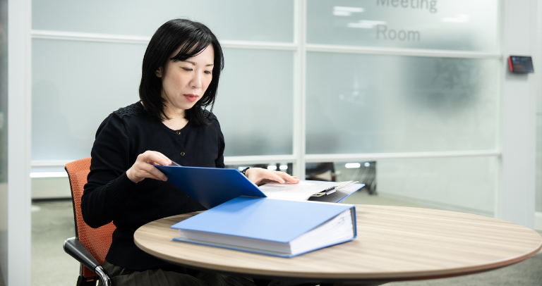 屋内の円卓に座り、書類の入った開いた青いフォルダーを手にしている人物。その人物の前の卓上には、他にもフォルダーが置かれている。