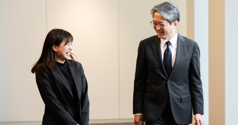 屋内で向かい合って立つ2人。フォーマルな服装を身に着け、背景は無地の壁。