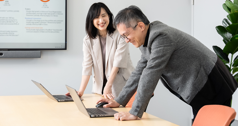 屋内の空間で、開いたノートパソコンを前にテーブルに身を乗り出す2人の人物がおり、背景には文字が表示されたスクリーンが見えている。