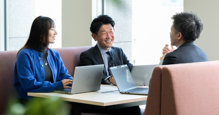 屋内のテーブルに3人が向かい合って座り、それぞれ開いたノートパソコンを前に、会話を交わしている。