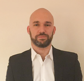 Individual wearing a white collared shirt and a dark suit jacket standing indoors against a plain, light-colored background in a formal portrait-style photograph.