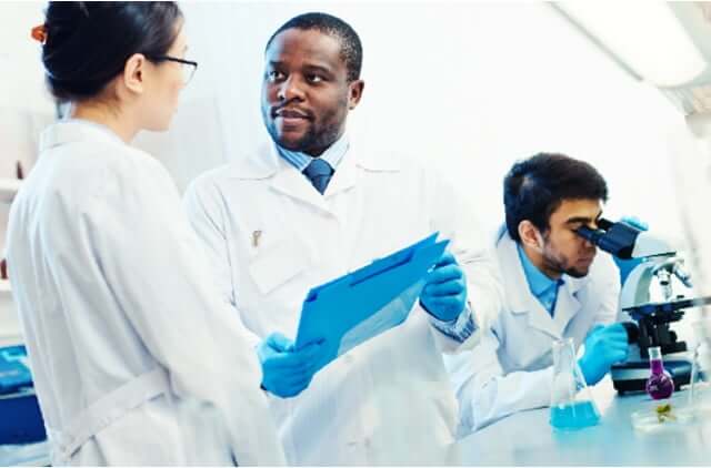 Two scientist in lab coats reviewing findings on a clipboard, while another scientist looks into a microscope