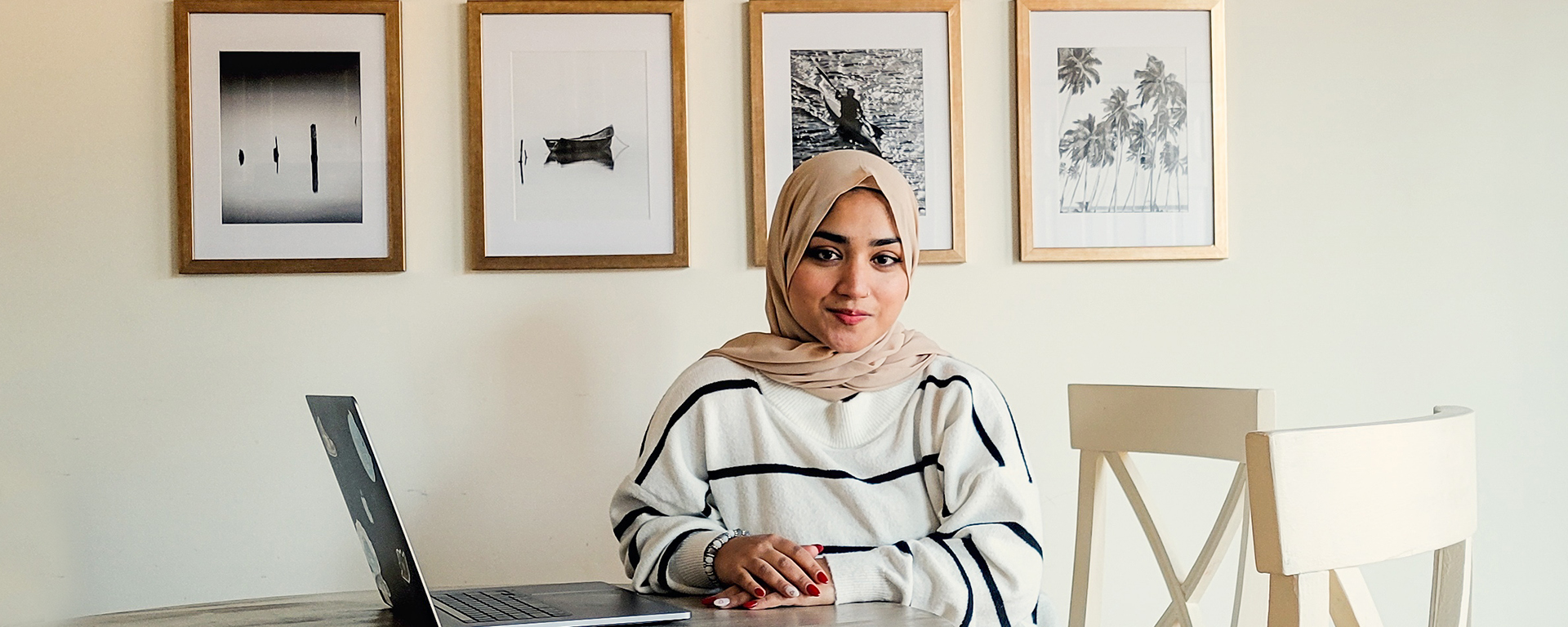 Capital One Cyber Security intern Khadija sits at her table with her laptop working from home behind a wall of picture frames