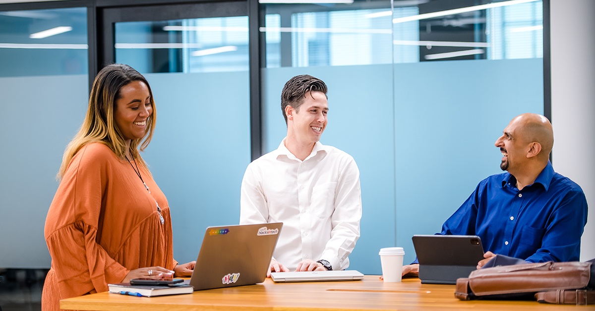 Three Capital One associates stand and sit around a table with their laptops in a meeting room