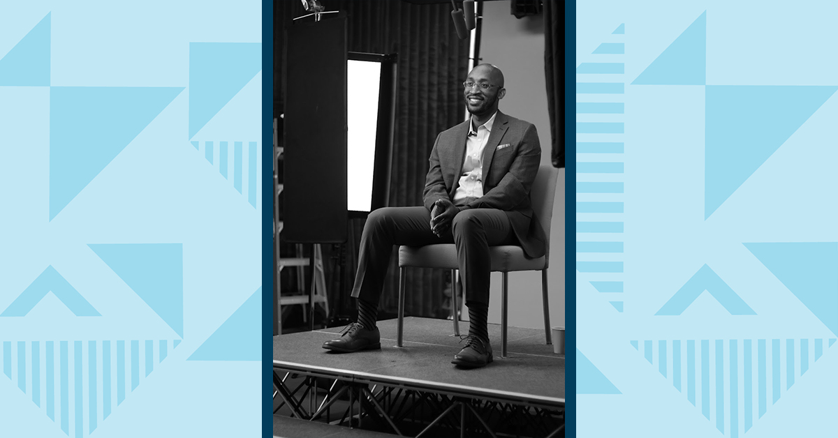 A black and white picture of Jerich, Capital One Tech associate, sitting in a chair in a suit for an interview, with a blue two-tone triangular background on either side of the picture