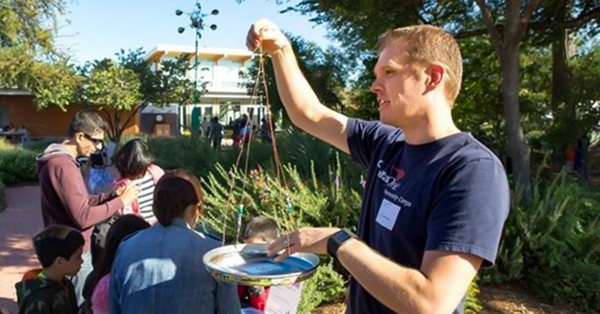 Capital One Green Teams Member Matt stands in a park next to people in front of trees and greenery and holds a birdfeeder