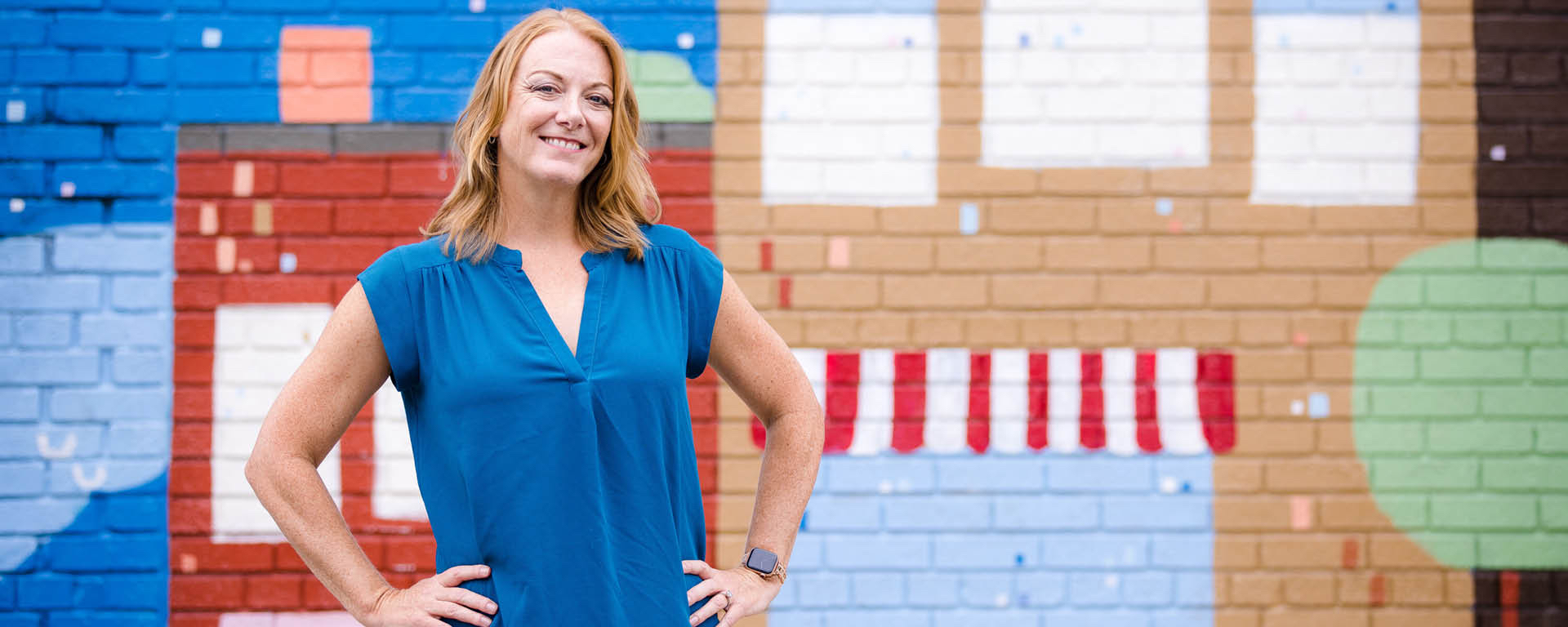 Heather Winkle, Capital One Senior VP & Head of Experience Design, stands and smiles in front of a brick wall with painted buildings on it