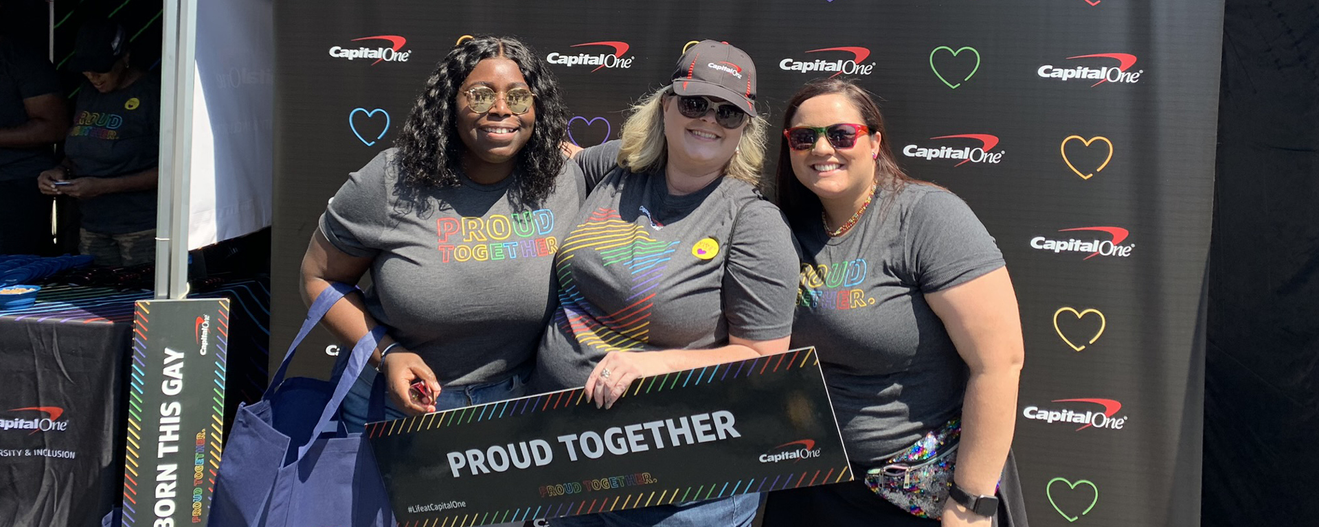 Three Capital One associates stand in front of a black backdrop with colorful hearts holding a sign that says 