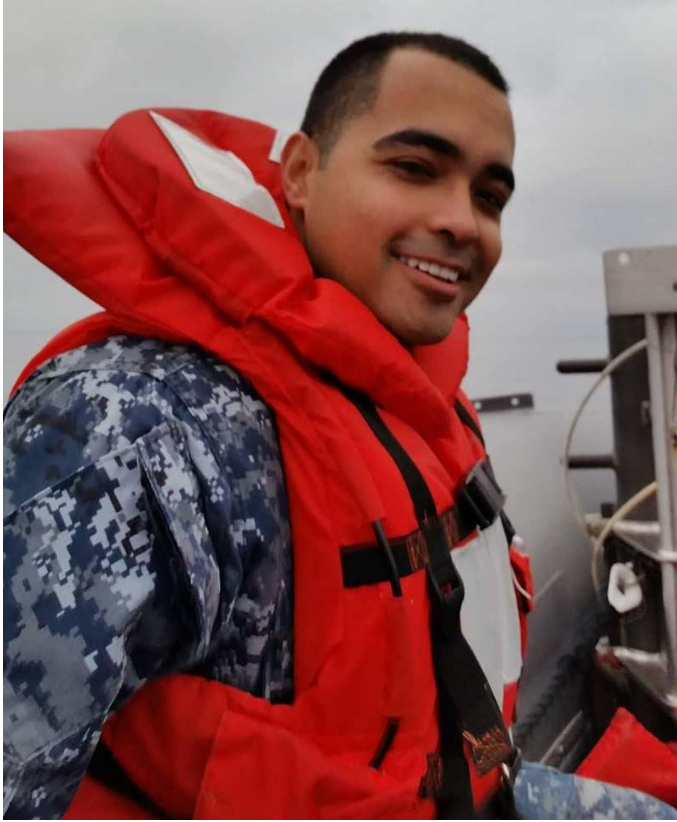 Photo of Alex wearing a military uniform and life vest, smiling to camera. 