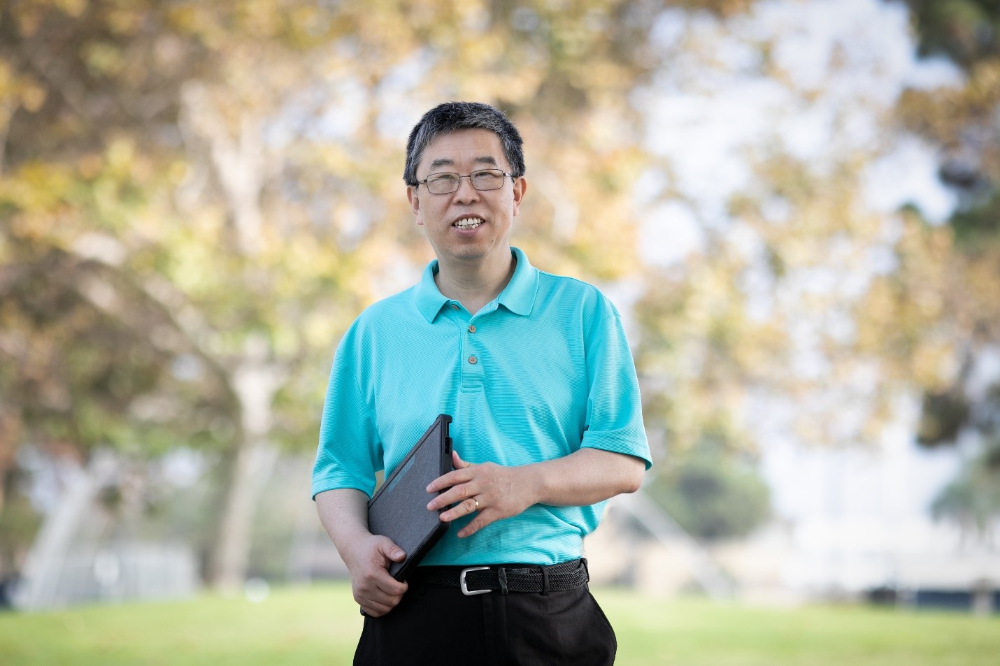 Image of Dr. Li standing outside in a park area, holding a laptop.