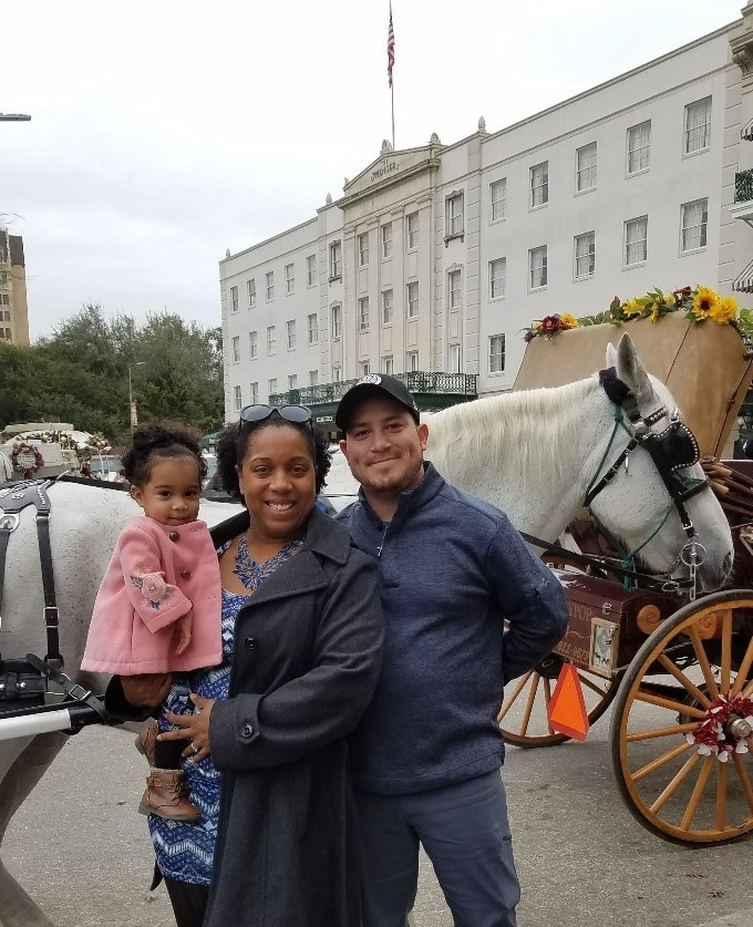 Aurelina is holding her baby daughter and standing beside her husband. They are stadning in front of a horse and carriage. 